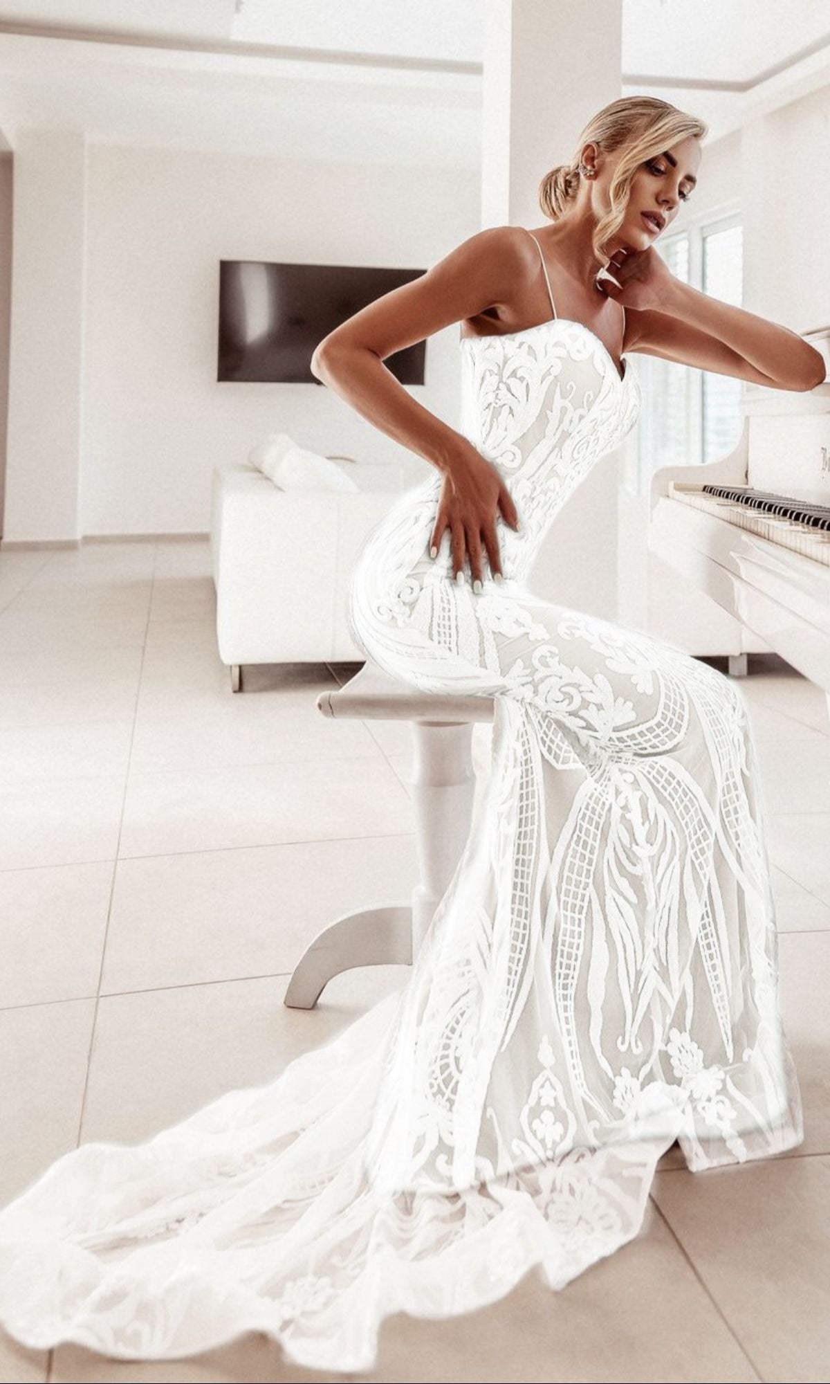A woman in a White Lace Bodycon Bridal Gown: Tina Holly BA999 sits on a white stool beside a piano in a bright, modern living room.